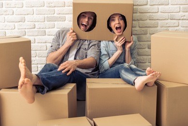 happy young couple is moving, hiding heads in boxes with holes, looking at camera and making faces, sitting among other cardboard boxes