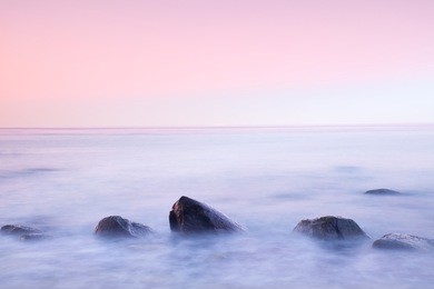 romantic atmosphere in peaceful morning at sea. big boulders sticking out from smooth wavy sea. pink horizon with first hot sun rays.