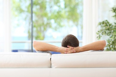rear view of a single man relaxed on a couch at home and looking the green background outdoors through the window in the livingroom