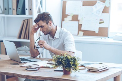 feeling tired. frustrated young handsome man looking exhausted while sitting at his working place and carrying his glasses in hand
