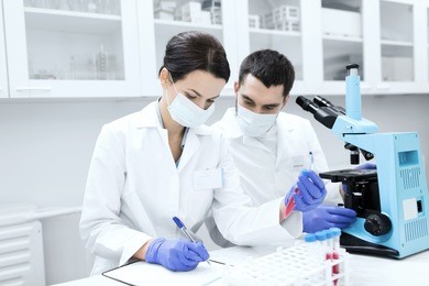 science, chemistry, technology, biology and people concept - young scientists with test tube and microscope making research in clinical laboratory and taking notes