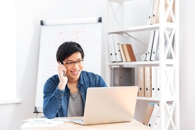 happy handsome young businessman in glasses using laptop and talking on cell phone in office
