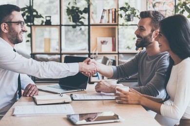 deal! side view of cheerful young man sitting close to his wife and shaking hand to man sitting in front of him at the desk 