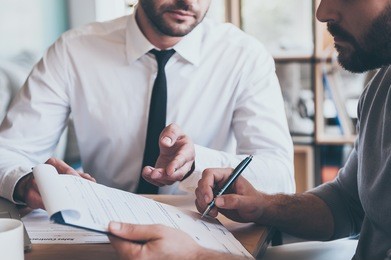 signing contract. close-up of confident young man signing some document while another man in shirt and tie sitting close to him and pointing document 