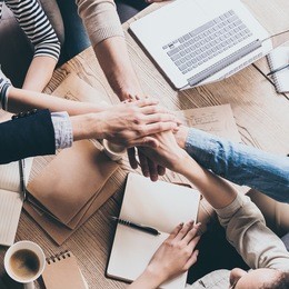 unity is a key to success. top view of people holding hands together while sitting at the wooden desk together 