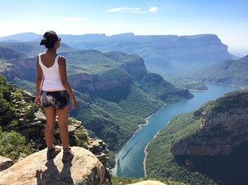 girl stands on a rock overlooking a canyon with a blue river in the bottom surrounded by green trees under a blue sky at the blyde river canyon in south africa.