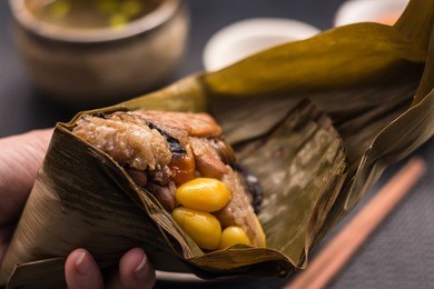freshly cooked zongzi wrapped with bamboo leaf holding and peeling on woman hands with dark background. zongzi is a traditional chinese food eaten during the dragon boat festival.