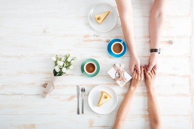 hands of young couple holding hands and drinking coffee on wooden table with cakes, gift box and flowers