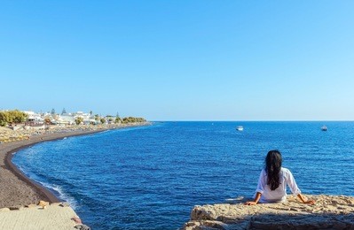 back of woman with black hair sitting on rock, enjoying peacefully the view of mediterranean sea