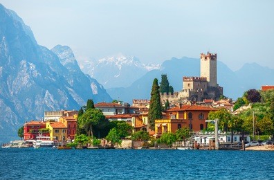 malcesine town. ancient tower and fortress in old town malcesine at garda lake, veneto region, italy. high snowbound top mountains on background summer landscape with colorful houses of malcesine bank