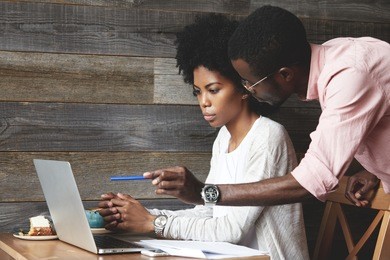 portrait of two colleagues working together at a cafe, discussing business ideas and plans, using laptop computer: african man holding a pen, pointing at the screen while presenting his project