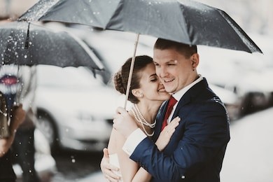 bride leans to groom's shoulder smiling while they walk across the street in rain