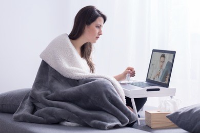 young woman covered with thick blanket, sitting on a bed in a bright room and having an on-line consultation with her doctor