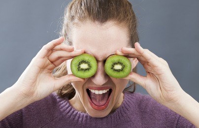 ecstatic beautiful young woman holding two cut kiwis on her eyes for fun joke or dynamic vitamin c, indoors