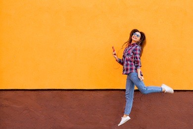 fashion portrait of pretty smiling and jumping woman in sunglasses with smartphone against the colorful orange wall. copyspace