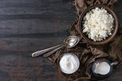 brown ceramic bowl of homemade cottage cheese with yogurt and jug of milk, served on wet sackcloth rag over dark textured iron background. healthy breakfast concept. top view with copy space.