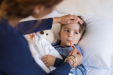 sick boy with thermometer laying in bed and mother hand taking temperature. mother checking temperature of her sick son who has thermometer in his mouth. sick child with fever and illness in bed.