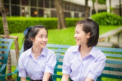 cute asian thai high schoolgirls student couple in school uniform chatting with her friend showing happy smile face expression on a bench  in vintage retro color