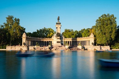 tourists and locals enjoy the summer heat rowing in the main pond and resting under the monument to alfonso xii at the retiro park (parque del buen retiro) in madrid, spain. long exposure.