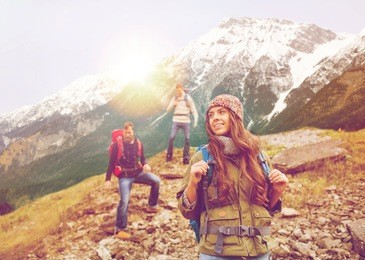 adventure, travel, tourism, hike and people concept - group of smiling friends with backpacks walking down downhill over alpine mountains and hills background