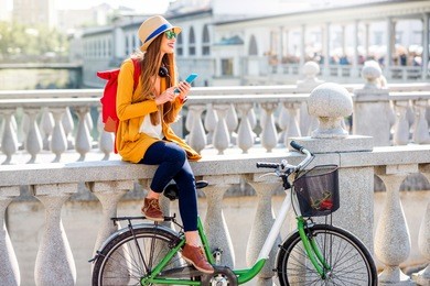 young female traveler sitting with bicycle and using smart phone on the triple bridge in ljubljana. traveling in slovenia