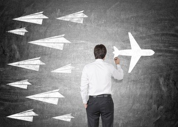 man in white shirt and black pants drawing sketch of modern airplane at blackboard. there are drawings of paper planes flying in different direction. concept of creativity and innovation.