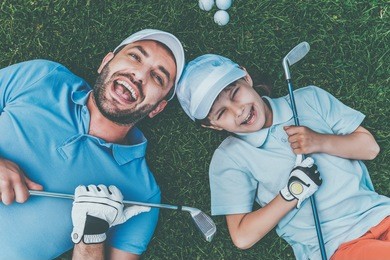 golf is fun! top view of cheerful little boy and his father holding golf clubs and smiling while lying on the green grass