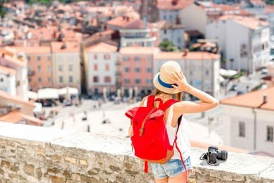 young female traveler with hat, red backpack and photo camera enjoying the view on piran old town. back view with copy space. traveling in slovenia