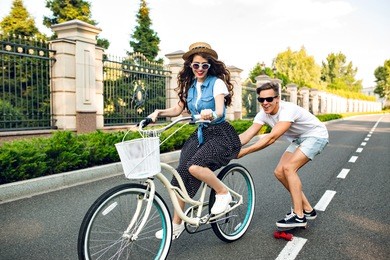 young couple having fun on summer on road. pretty girl with long hair driving a bike, handsome guy  keeps for a bicycle and rides on a skateboard. they look exited