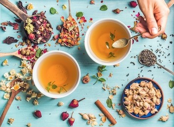 two cups of healthy herbal tea with mint, cinnamon, dried rose and camomile flowers in spoons and man's hand holding spoon of honey, blue background, top view