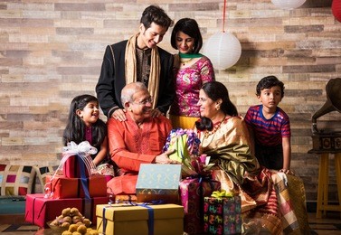 3 generations of indian family celebrating diwali festival or birthday by exchanging gifts and sweets while sitting on sofa, indoors