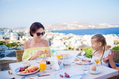 family having breakfast at outdoor cafe with amazing view on mykonos town. adorable girl and mom drinking fresh juice and eating croissant on luxury hotel terrace