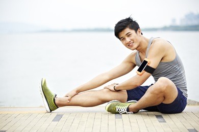 young handsome asian jogger wearing fitness tracker stretching a leg by lake looking at camera smiling