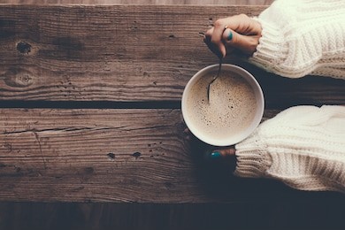 woman holding cup of hot coffee on rustic wooden table, closeup photo of hands in warm sweater with mug, winter morning concept, top view