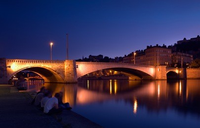 llluminated bridge over the river saone in the city of lyon, france, with a group of young people  having a friendly chat by the river