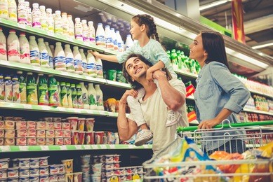 beautiful young parents and their cute little daughter are smiling while choosing food in the supermarket. girl is sitting on her dad's shoulders