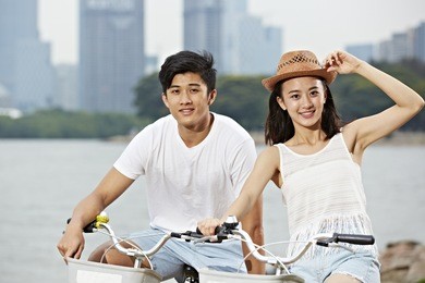 young asian couple riding bike in urban park, looking at camera smiling
