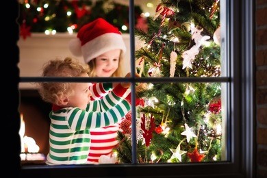 happy little kids in matching red and green striped pajamas decorate christmas tree in beautiful living room with traditional fire place. children opening presents on xmas eve. view though window.