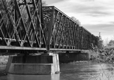 a black and white landscape photo of a railroad bridge on the american countryside. a river flows underneath the bridge.