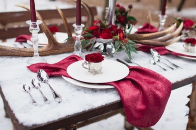 wedding table setting in marsala colors with plates, cutlery, red floral compositions, candles, velvet napkins, deer horns on table covered with snow