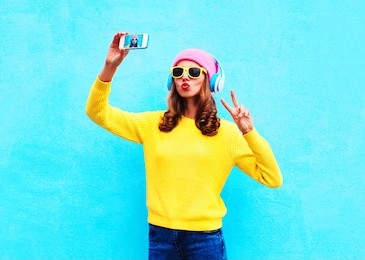 fashion cool girl in headphones listening music taking photo makes self portrait on smartphone wearing a colorful clothes over blue background