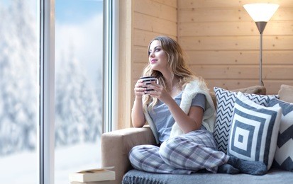 young beautiful blonde woman with cup of coffee sitting home in living room by the window. winter snow landscape view. lazy day off concept