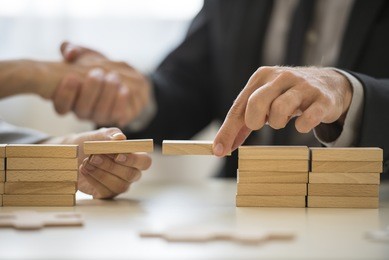 teamwork or building bridges concept with a businessman and woman holding wooden building blocks to form a bridge over a gap while clasping hands in the background.