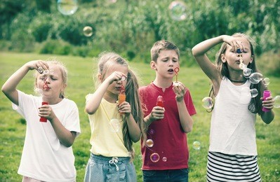 cheerful boy and girls in elementary school age standing on green grass and blowing bubbles 