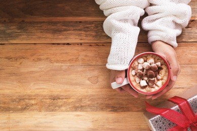 female hand holding cup of hot cocoa or chocolate with marshmallow on wooden table from above
