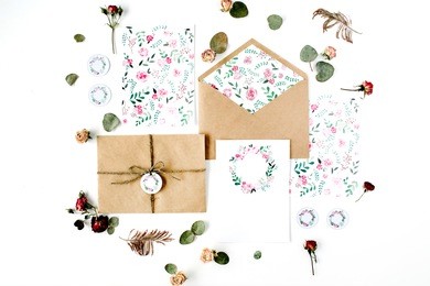 flat lay workspace. wedding invitation cards, craft envelopes, pink and red roses and green leaves on white background. overhead view, top view
