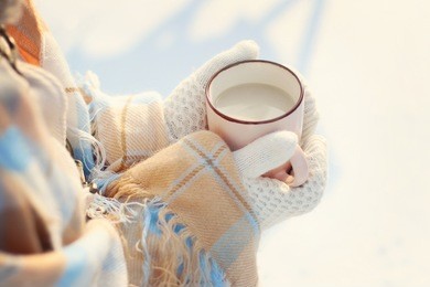 girl covered with plaid and in white knitted mittens holding in hands hot cocoa, tea with milk outdoors on snowy background. sweet cozy winter time. winter wonderland. winter still life
