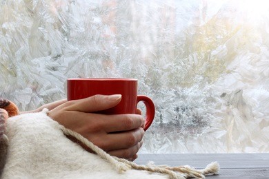 big red mug in the hands thrown over with a warm blanket against the window with frost / warming drink at home in winter