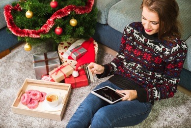 young smiling woman wearing sweater and jeans is buying in internet with credit card and tablet. christmas tree, tea, sweets and gifts on background. christmas and new year time. internet shopping