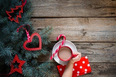 child hand holds a cup of cocoa drink in red cup. christmas tree with decor ,  pink candy cane, red napkin at white polka dots pattern on wooden table, top view